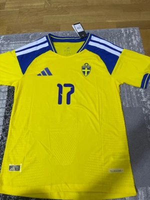 Sverige fotbollströja Gyökeres #17 - Gul och blå officiell Sverige fotbollströja från Adidas med Gyökeres och nummer 17 på ryggen. Tröjan har blå detaljer, landslagets emblem på bröstet och CLIMACOOL-material som andas. Perfekt för match eller träning.