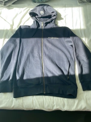 Sail Racing hoodie mörk blå - Snygg marinblå hoodie från Sail Racing med dragkedja framtill och två fickor med dragkedja. Klassisk logga på bröstet och huva med dragsko. Tillverkad i mjukt bomullsmaterial som känns skönt mot huden. Perfekt för en avslappnad och sportig stil.
