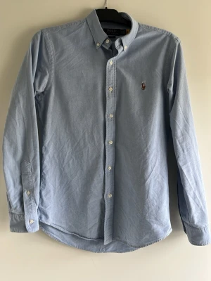 Ljusblå skjorta från Polo Ralph Lauren - Klassisk ljusblå skjorta från Polo Ralph Lauren med button-down krage och broderad logga på bröstet. Skjortan har långa ärmar, vita knappar och är tillverkad i mjuk bomull. Perfekt för en clean och stilren look.