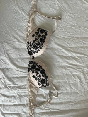 Intimissimi beige push-up bh med svart spets - Snygg beige push-up bh med vadderade kupor och svart blommig spetsbrodyr framtill. Axelbanden är justerbara och bh:n har spetsdetaljer längs kanterna för en extra feminin touch. Perfekt för dig som gillar romantiska detaljer.