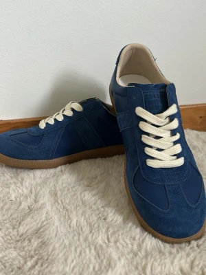 Blå sneakers från Maison Margiela - Snygga blå sneakers från Maison Margiela med ovandel i mocka och textil, vita snören. Skorna har en snygg design. Riktigt snygga till sommaren. Stl 41 och de med följer även en dustbag. Pris kan diskuteras vid snabb affär, har ni någon fråga är de bara att skriva.