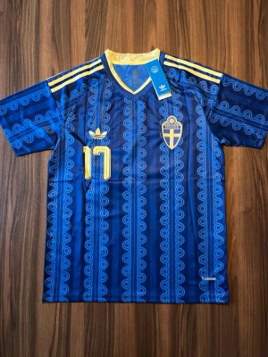 Blå Sverige fotbollströja Adidas - Säljer en blå fotbollströja från Adidas med Sveriges landslagsemblem och nummer 17 Gyökeres på ryggen. Tröjan har gula detaljer, korta ärmar och ett unikt mönster i blå nyanser. Klassiska Adidas-ränder på axlarna och V-ringad krage.