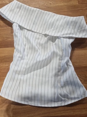 Vit oneshoulder randig topp - Snygg vit one shoulder topp med tunna svarta ränder. Den har en drag kedja på sidan