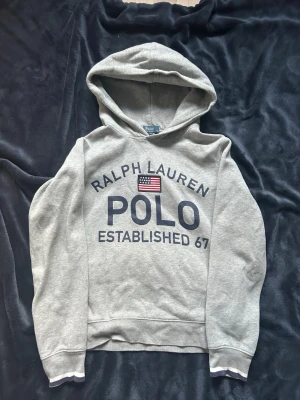 Grå hoodie från Polo Ralph Lauren - Snygg grå hoodie från Polo Ralph Lauren med stor logga och amerikansk flagga broderad på bröstet. Hoodien har huva och ribbade muddar vid ärmslut och nederkant. Tillverkad i mjukt bomullsmaterial, perfekt för en avslappnad och trendig look.
