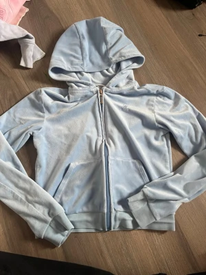 Ljusblå velourhoodie Juicy Couture - Säljer en ljusblå hoodie i mjuk velour från Juicy Couture. Hoodien har dragkedja framtill, två fickor och en broderad logga på bröstet. Perfekt för en chill och trendig look. Klassisk huva och ribbade muddar vid ärmar och nederkant.