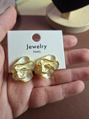 Blommiga örhängen i guld och pärlemor - Unika örhängen i form av blommor med skimrande pärlemor och guldfärgade kanter. De har en modern, konstnärlig design som verkligen sticker ut och ger en lyxig känsla. Perfekta för dig som vill ha något extra till din stil.