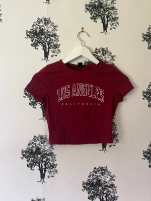 Röd croppad t-shirt Los Angeles - Snygg röd croppad t-shirt med trycket 'Los Angeles California' framtill. T-shirten har korta ärmar och rund halsringning. Perfekt för en avslappnad och trendig stil.