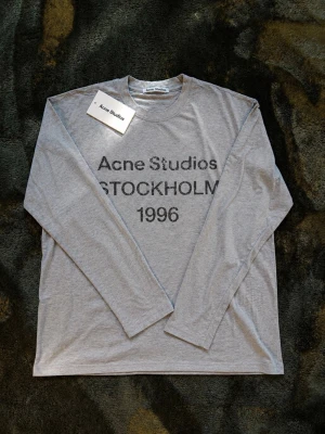 Grå långärmad topp Acne Studios - Snygg grå långärmad topp från Acne Studios med tryck 'STOCKHOLM 1996' framtill. Tillverkad i mjuk bomull med avslappnad passform och rund halsringning. Perfekt för en clean och stilren look. Den ser lite ljusare ut på bilderna än vad de är igentligen vet inte vad de beror på dock. 