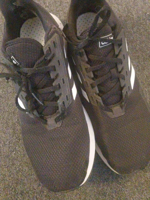Svarta Adidas sneakers med vita ränder - Snygga svarta sneakers från Adidas med klassiska vita ränder på sidorna. Ovandelen är i meshmaterial för bra andningsförmåga och sulan är vit. Skorna har svarta snören och en sportig look som passar till många outfits.