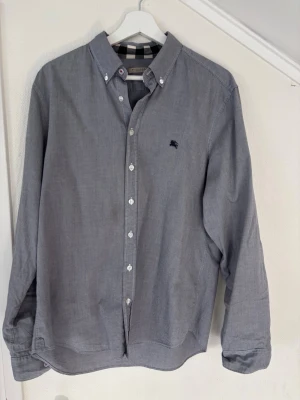 Burberry Skjorta - Klassisk grå skjorta från Burberry Brit med button-down krage och vita knappar. Skjortan har lång ärm och diskret Burberry-logga broderad på bröstet. Tillverkad i mjuk bomull och har det ikoniska Burberry-mönstret på insidan av kragen.           Köptes inför förra sommaren men användes enbart ett fåtal gånger. Kan givetvis lösa fler bilder vid önskemål. Kolla gärna på liknande annonser under min profil. Uppskattar snabba affärer 🙏