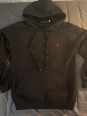 Svart hoodie från Polo Ralph Lauren - Snygg svart hoodie från Polo Ralph Lauren med klassisk röd logga broderad på bröstet. Hoodien har huva med dragsko och en stor magficka framtill. Tillverkad i mjukt material som känns skönt mot huden. Perfekt för en avslappnad och stilren look.
