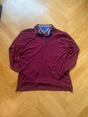 Vinröd långärmad Henley från Tommy Hilfiger - Snygg vinröd långärmad Henley från Tommy Hilfiger med klassisk krage och diskret logga på bröstet. Skjortan har knappar vid halsen och kontrastfärgade detaljer på insidan av kragen. Tillverkad i mjuk bomull för en bekväm känsla.