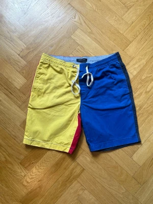 Färgglada colorblock shorts med snörning - Säljer ett par unika colorblock shorts med snörning i midjan. Framsidan är gul och blå med en röd detalj, baksidan är röd och mörkblå. Perfekta för sommaren och riktigt sköna att bära. Shorts i bomull med elastisk midja och fickor bak.