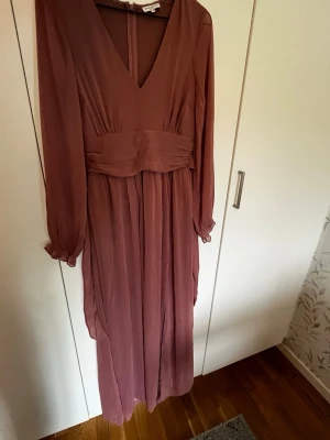 Lång rosa klänning med v-ringning - Säljer en elegant långklänning i typ dusty pink med v-ringad front och markerad midja. Klänningen har långa ärmar med lätt puff. Från bubbleroom storlek xs. Aldrig använd. Dragkedjan krånglar ibland men man får bara ta i lite så åker den upp. 