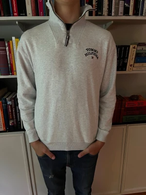 Grå halvzip från Tommy Hilfiger - Snygg ljusgrå halvzip-tröja från Tommy Hilfiger med hög krage och dragkedja. Diskret logga på bröstet och liten flagga på ärmen. Tillverkad i mjukt bomullsmaterial och har ribbade muddar vid ärmslut och nederkant. Perfekt för en avslappnad och stilren look. Stl164/XS men passar som S. Modellen är 179 och väger 65kg.