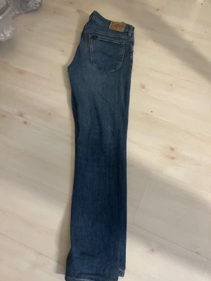 Blå raka jeans från Lee - Säljer ett par klassiska blå jeans från Lee med rak passform och femficksmodell. Jeansen har en mörkblå tvätt och detaljer i brunt läder på bakfickan. Perfekta för en avslappnad stil. Det är storlek 30/30.