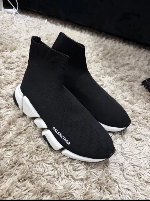 Balenciaga Speed Trainer svarta sneakers - Svarta Balenciaga Speed Trainer sneakers med vit chunky sula och stickad strumpliknande ovandel. Minimalistisk design med Balenciaga-logga på sidan. Skorna har en elastisk passform och är tillverkade i textil och gummi.