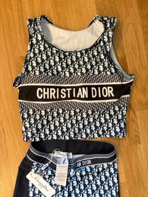  Träning sett med logotypmönster - Snyggt tvådelat set med svartvitt logotypmönster. Setet består av ett ärmlöst crop top-linne och matchande shorts med bred resår i midjan. Tydlig Christian Dior-text på både linne och shorts. Tillverkat i Frankrike. Oanvänt . 