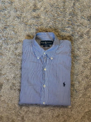 Blårutig skjorta från Ralph Lauren - Klassisk blårutig skjorta från Ralph Lauren med button-down krage och broderad logga på bröstet. Skjortan har långa ärmar, vita knappar och är tillverkad i mjuk bomull. Perfekt för en stilren och avslappnad look.
