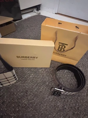Burberry bundle rutigt bälte med logga + en plånbok på köpet - Snyggt bälte från Burberry med klassiskt rutigt mönster i beige och svart. Bältet har en stilren metallspänne med Burberrys logga framtill. Perfekt accessoar för att lyfta din outfit.