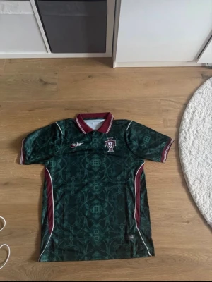 Grön Portugal fotbollströja Nike - Snygg grön Portugal fotbollströja från Nike med mönstrad design, röd-vit krage och detaljer längs sidorna. Tröjan har korta ärmar och Portugal-emblem på bröstet. Perfekt för dig som vill sticka ut på planen eller heja på landslaget.