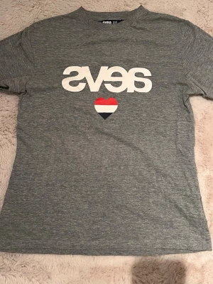 Grå t-shirt från Svea med tryck - Snygg gråmelerad t-shirt från Svea med stort vitt tryck och ett hjärta i rött, vitt och blått på bröstet. Klassisk passform och rund halsringning. Perfekt till jeans eller shorts för en avslappnad look.