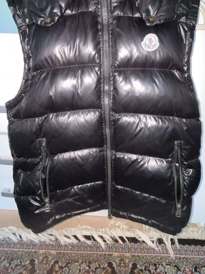 Svart dunväst från Moncler - Snygg svart dunväst från Moncler med glansig finish och huva. Västen har två stora fickor med dragkedja framtill och Moncler-logga på bröstet. Perfekt för lager-på-lager och håller dig varm under kyliga dagar.