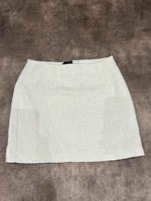 Vit kortkjol i bouclé med dragkedja - Stilren vit kortkjol i bouclé-liknande material med diskret struktur. Kjolen har en smal passform och stängs med en dold dragkedja i sidan. Perfekt att matcha med en enkel topp eller kavaj för en trendig look.