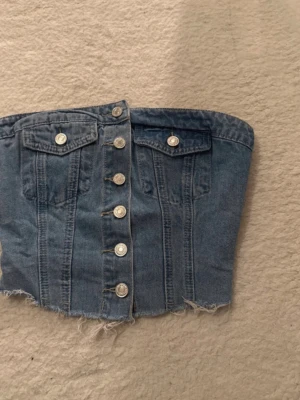 Blå denim korsett-tubtopp med knappar - Trendig blå denim korsett-tubtopp med knappar framtill och råa kanter nertill. Toppen har två fejkfickor med knappdetaljer och smockad baksida för extra stretch. Perfekt för en edgy och cool look. Använd endast 1 gång så jätte bra skick.
