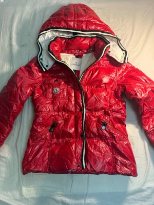 Röd dunjacka med huva från Moncler - Snygg röd dunjacka från Moncler med glansig finish och avtagbar huva. Jackan har svarta och vita detaljer vid dragkedjan, flera fickor och klassisk Moncler-logga på bröstet. Insidan har ett unikt serietryck. Perfekt för kalla vinterdagar.ej äkta
