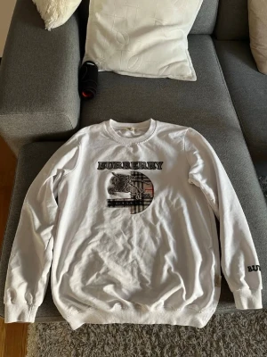 Burberry sweatshirt - Vit stickad tröja från Burberry med broderad logga och klassiskt rutmönster på bröstet. Tröjan har rund halsringning, långa ärmar och ribbade muddar. Snygg detalj med BURBERRY-tryck även på ärmen. Kan tänka går ner i pris via direkt köp