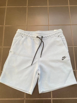 Ljusblå Nike shorts med snörning - Säljer ett par ljusblå shorts från Nike med svart snörning i midjan och diskret Nike-logga på vänster ben. Shortsen har fickor fram och en bakficka med svart dragkedja. Perfekta för sommaren och riktigt sköna att bära.