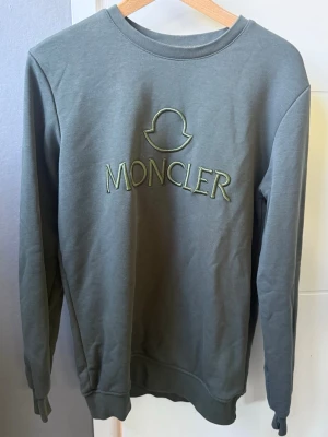 Grön sweatshirt från Moncler - Stilren grön sweatshirt från Moncler med broderad logotyp och text på bröstet. Tröjan har rund halsringning, långa ärmar och ribbade muddar vid ärmslut och nederkant. Äkta givetvis. Storlek M (true to size). Mycket fint skick! Condition: 8/10 