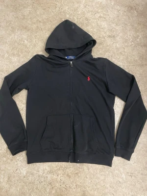 Svart hoodie från Polo Ralph Lauren - Klassisk svart hoodie från Polo Ralph Lauren med dragkedja framtill och röd broderad logga på bröstet. Hoodien har huva, två fickor fram och ribbade muddar vid ärmslut och nederkant. Tillverkad i mjukt bomullsmaterial för en skön känsla.