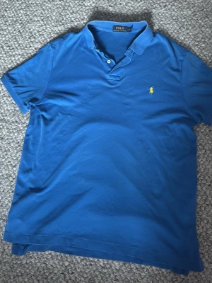 Blå piké från Polo Ralph Lauren - Klassisk blå pikétröja från Polo Ralph Lauren med gul broderad logga på bröstet. Tröjan har krage, korta ärmar och knappar vid halsen. Perfekt till jeans eller shorts för en avslappnad men snygg look.