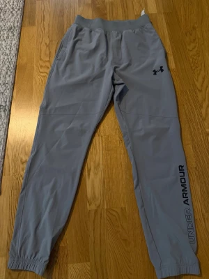 Grå mjukisbyxor från Under Armour - Snygga grå mjukisbyxor från Under Armour med resår i midjan och benslut. Byxorna har dragsko i midjan, två sidofickor och logga på låret samt text längs benet. Tillverkade i ett mjukt och stretchigt material som känns skönt mot huden.