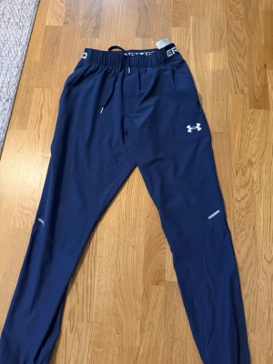 Blå träningsbyxor från Under Armour - Mörkblå träningsbyxor från Under Armour med resår och dragsko i midjan. Byxorna har vita logodetaljer, reflexdetaljer på benen och är tillverkade i ett lätt, snabbtorkande material. Perfekta för löpning eller gym.