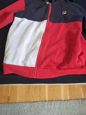 FILA hoodie i rött, vitt och svart - Snygg hoodie från FILA i färgblock med rött, vitt och svart. Dragkedja framtill, huva och liten FILA-logga på bröstet. Tillverkad i mjukt material, perfekt för en avslappnad streetwear-look.