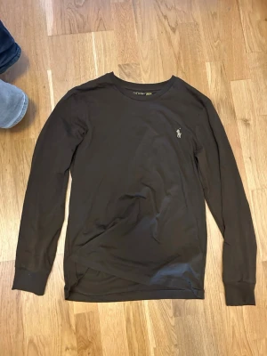 Brun långärmad topp från Polo Ralph Lauren - Klassisk brun långärmad topp från Polo Ralph Lauren med den ikoniska broderade loggan på bröstet. Tillverkad i mjuk bomull som känns skön mot huden. Enkel och stilren design som funkar till det mesta.