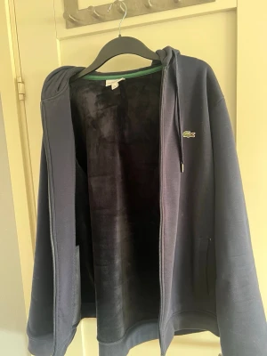 Mörkblå hoodie från Lacoste - Mörkblå hoodie från Lacoste med dragkedja, mjukt foder och klassisk krokodil-logga på bröstet. Stor huva med texten 'LACOSTE' tryckt i vitt på baksidan. Två fickor framtill och ribbade muddar. Perfekt för en chill och sportig look. Speciell material på insidan så jätte bra till vintern
