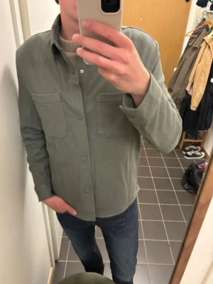 Grön overshirt från Zara - Stilren grön overshirt från Zara med två stora bröstfickor och knappar framtill. Skjortjackan har en rak passform och är tillverkad i ett mjukt tyg som passar perfekt för lager på lager. Enkel att matcha med jeans eller chinos.