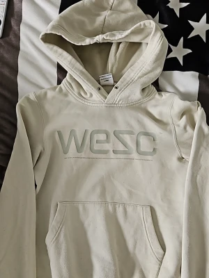 Beige hoodie från WESC med logga - Säljer en beige hoodie från WESC med stor logga på bröstet. Hoodien har en klassisk huva med snörning och en stor magficka. Materialet är mjukt och bekvämt, perfekt för chill dagar. Långärmad och enkel design som passar till det mesta.