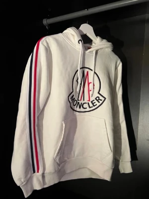 Vit Moncler hoodie med ränder - Snygg vit hoodie från Moncler med stor logga på bröstet och röda, vita och blå ränder längs ärmarna. Klassisk huva med dragsko och känguruficka framtill. Moncler-märke på ena ärmen. Perfekt för dig som gillar streetwear och exklusiva märken.