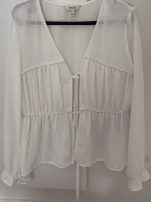 Vit blus med knytband från Vero Moda - Stilren vit blus från kollektionen AWARE med v-ringning och knytband. Blusen har långa ärmar med resår vid ärmsluten och ett lätt rynkat fall. Tillverkad i ett tunt, strukturerat tyg som känns luftigt.