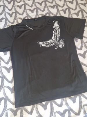 Svart t-shirt med örntryck - Cool svart t-shirt med ett stort vitt örntryck på bröstet. Klassisk rund halsringning och korta ärmar. Perfekt för dig som gillar streetwear och vill sticka ut med en unik design. Inga defekter