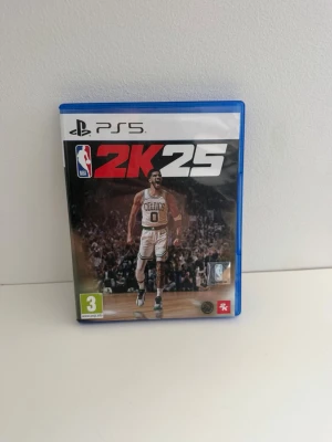 NBA 2K25 PS5 spel - NBA 2K25 till PlayStation 5 med blå plastfodral och omslag som visar en basketspelare från Celtics. Spelet är ett sportspel med baskettema och inkluderar en pre-order bonus. Skivan har röd och svart design med PS5-logga. Använd men i mycket gott skick