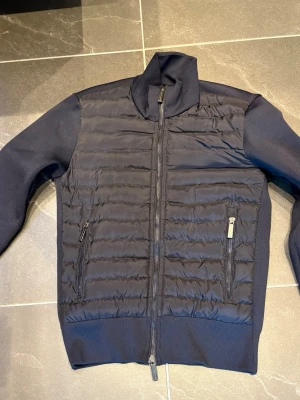 Mörkblå cardiganjacka från Canada Goose - Snygg mörkblå cardiganjacka från Canada Goose med quiltad front och stickade ärmar. Jackan har två dragkedjeförsedda fickor fram och en hög krage. Klassisk Canada Goose-logga på ärmen. Perfekt för dig som vill ha en stilren och bekväm jacka.