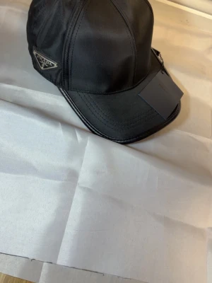 Svart keps från Prada  - Snygg svart keps från Frada Milano med klassisk böjd skärm och ett metallmärke på sidan. Kepsen är tillverkad i ett glansigt syntetmaterial och har tydliga sömmar som ger en sportig look. Perfekt accessoar för en trendig streetstyle.