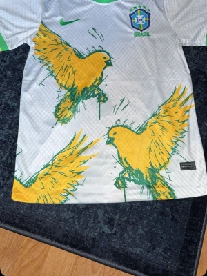 Brasilien Nike matchtröja papegojmotiv - Snygg vit Brasilien matchtröja från Nike med stora gula papegojor i grönt och gult tryck. Tröjan har gröna detaljer vid ärmslut och krage samt Brasilien-emblem och Nike-logga på bröstet. Perfekt för dig som älskar fotboll och vill sticka ut på planen. Helt ny aldrig andvänd med påse och tags