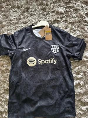 Svart FC Barcelona Nike matchtröja - Snygg svart FC Barcelona matchtröja från Nike med diskret paisleymönster, vit Nike-logga och klubbmärke på bröstet samt Spotify som sponsor. Tillverkad i lätt och ventilerande Dri-FIT material, perfekt för träning eller match.